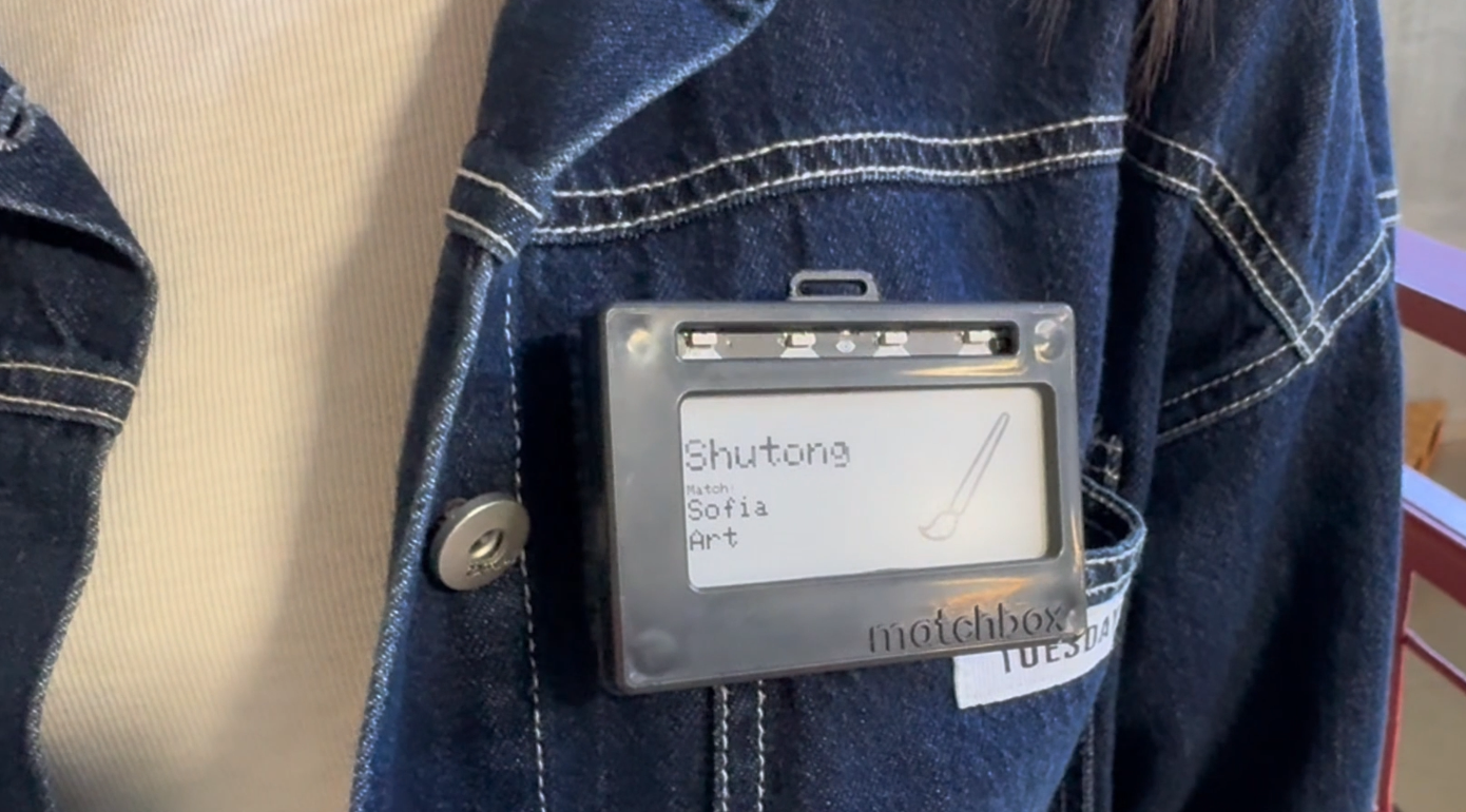 Matchbox badge attached to a denim jacket with magnetic standoffs, e-ink display showing a name, match, and topic icon